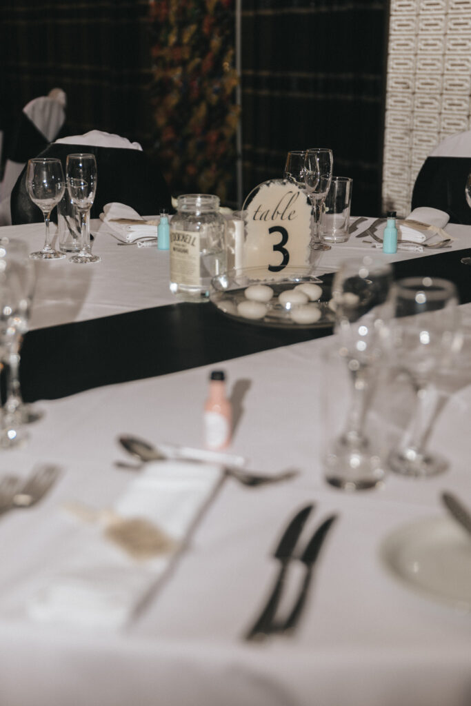 Elegant table setting at the Brackenborough Hotel with a white tablecloth and a black runner. Features cutlery, glassware, and folded napkins. A jar, small bottles, and a dome with "Table 3" are central. Blurred background includes a decorative wall and foliage, perfect for any wedding photographer's portfolio. © Aimee Lince Photography - Wedding photographer in Lincolnshire, Yorkshire & Nottinghamshire