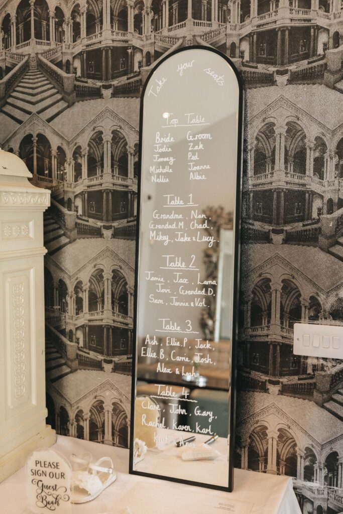 A full-length mirror at the Brackenborough Hotel Wedding showcases a seating chart, elegantly displaying five tables labeled with names. The vintage architectural wallpaper serves as the perfect backdrop. Below, a sign on a cabinet invites guests to "Please sign our guest book," accompanied by a pen and glass heart. © Aimee Lince Photography