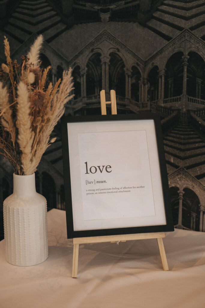 A framed definition of "love" on a small easel graces the table, symbolizing the essence of marriage. Nearby, a white vase holds dried pampas grass against a black-and-white architectural pattern. The definition reads: "A strong and passionate feeling of affection for another person or intense emotional attachment. © Aimee Lince Photography