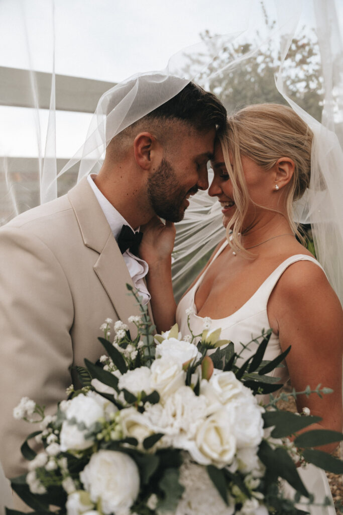A bride and groom gently touch foreheads, smiling under the sheer fabric of a veil. Captured in a photo at the Brackenborough Hotel, the groom wears a light beige suit with a black bow tie, while the bride holds a bouquet of white flowers and greenery. The softly blurred background hints at an outdoor marriage setting. © Aimee Lince Photography