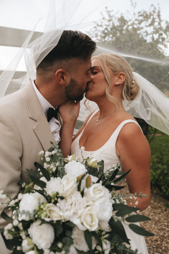 A bride and groom share a tender kiss under a delicate veil, embodying the essence of marriage. The bride, holding a bouquet of white roses and greenery, wears a sleeveless white gown with her hair in an updo. The groom stands elegantly in a beige suit with a black bow tie amidst the lush backdrop of their wedding scene. © Aimee Lince Photography