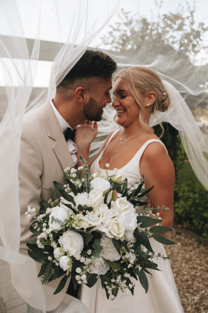 A bride and groom stand close under a veil, smiling at each other during their Brackenborough Hotel wedding. The bride holds a bouquet of white roses and greenery, wearing a white sleeveless gown. He wears a beige suit with a black bow tie, set against greenery and subtle outdoor lighting. © Aimee Lince Photography