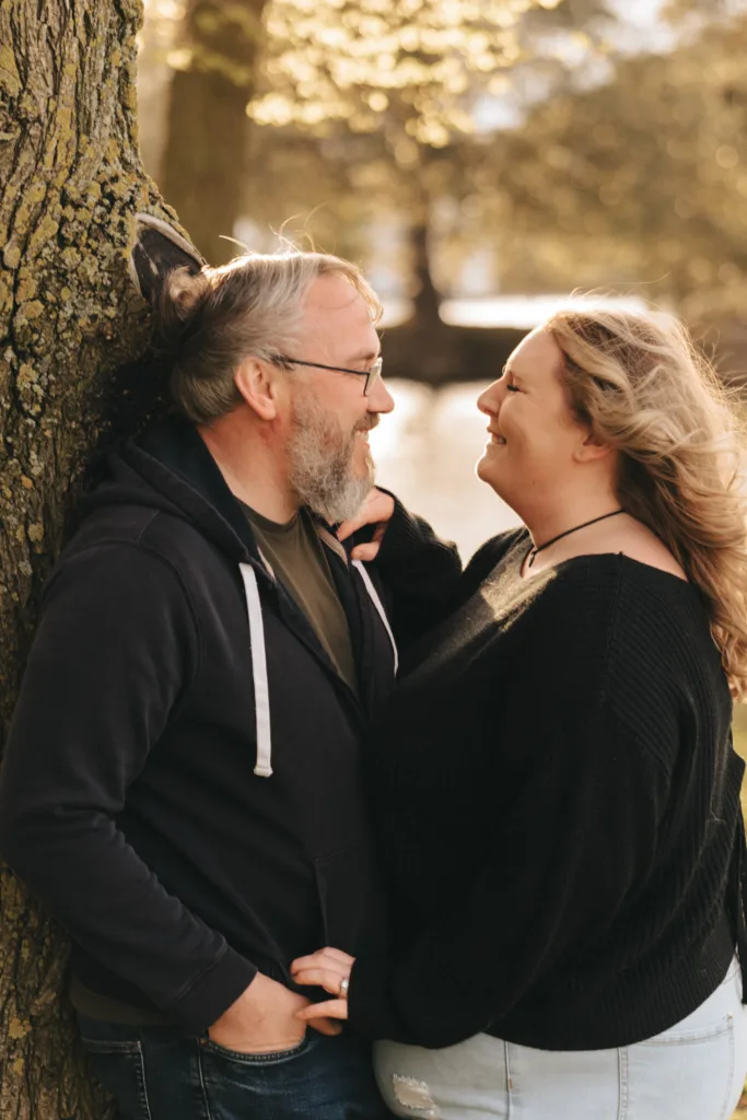 A smiling couple stands close together outdoors by a tree at Cleethorpes Boating Lake. The man, with a grey beard and glasses, wears a black hoodie and jeans. The woman, with long blonde hair and a black sweater, leans towards him. Sunlight filters through the trees, creating an engagement-like warmth. © Aimee Lince Photography - Wedding photographer in Lincolnshire, Yorkshire & Nottinghamshire