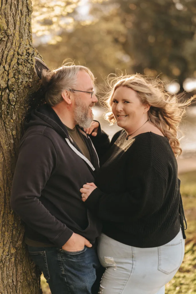 A man with glasses and a beard, wearing a dark hoodie, leans against a tree while smiling at a woman with wavy hair in a black sweater and ripped jeans. They stand close by Cleethorpes Boating Lake in the sunlit park, with trees and water blurred behind them, both looking happy and relaxed. © Aimee Lince Photography - Wedding photographer in Lincolnshire, Yorkshire & Nottinghamshire