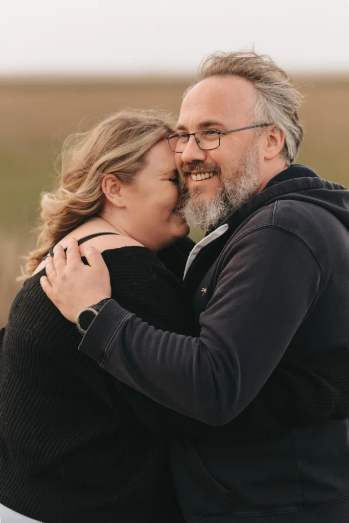 A couple embraces outdoors, both smiling, as if celebrating an engagement. The woman leans her head against the man's shoulder, eyes closed. The man, wearing glasses and a beard, complements her dark sweater. The blurred background, reminiscent of Cleethorpes Boating Lake, features earthy tones. © Aimee Lince Photography - Wedding photographer in Lincolnshire, Yorkshire & Nottinghamshire
