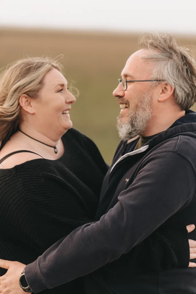 A smiling couple stands outdoors in a close embrace, hinting at their engagement. The woman, with long wavy hair, wears a black top and necklace. The bearded man sports glasses and a dark hoodie. Behind them is the blurred landscape of Cleethorpes Boating Lake, enhancing the romantic setting. © Aimee Lince Photography - Wedding photographer in Lincolnshire, Yorkshire & Nottinghamshire