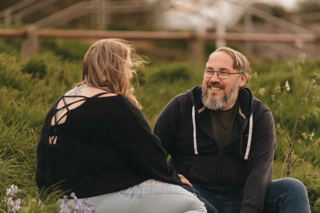 A woman with long blonde hair and a man with a beard sit on the grass near Cleethorpes Boating Lake, smiling and engaged in conversation. The woman sports a black top with crisscross back design, while the man wears glasses, a dark hoodie, and jeans. They are outdoors near a blurred fence surrounded by greenery. © Aimee Lince Photography - Wedding photographer in Lincolnshire, Yorkshire & Nottinghamshire