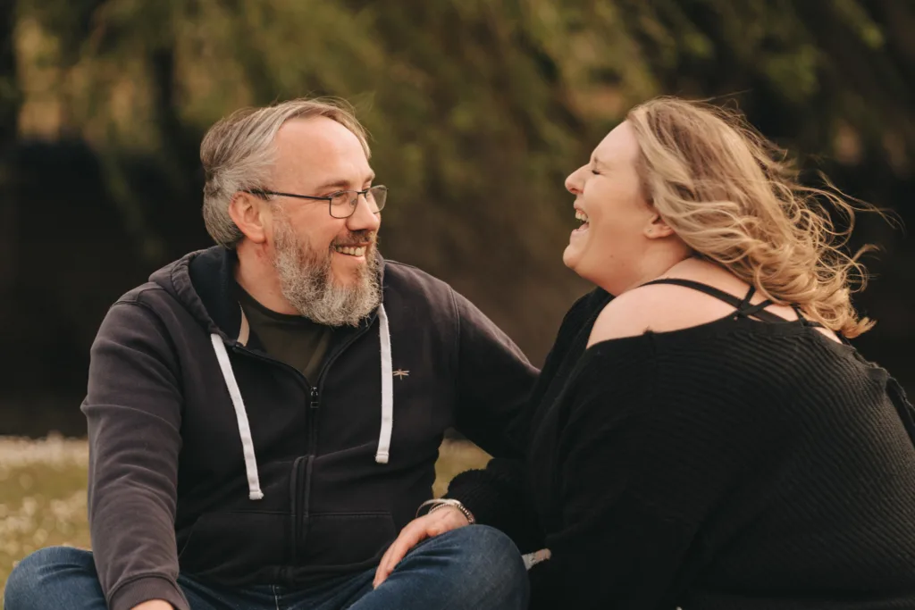 A man with glasses and a beard, in a black hoodie, sits on the grass near Cleethorpes Boating Lake with a woman. Her windblown hair complements her black sweater as they laugh together, sharing a moment of engagement amidst the blurred greenery backdrop. © Aimee Lince Photography - Wedding photographer in Lincolnshire, Yorkshire & Nottinghamshire
