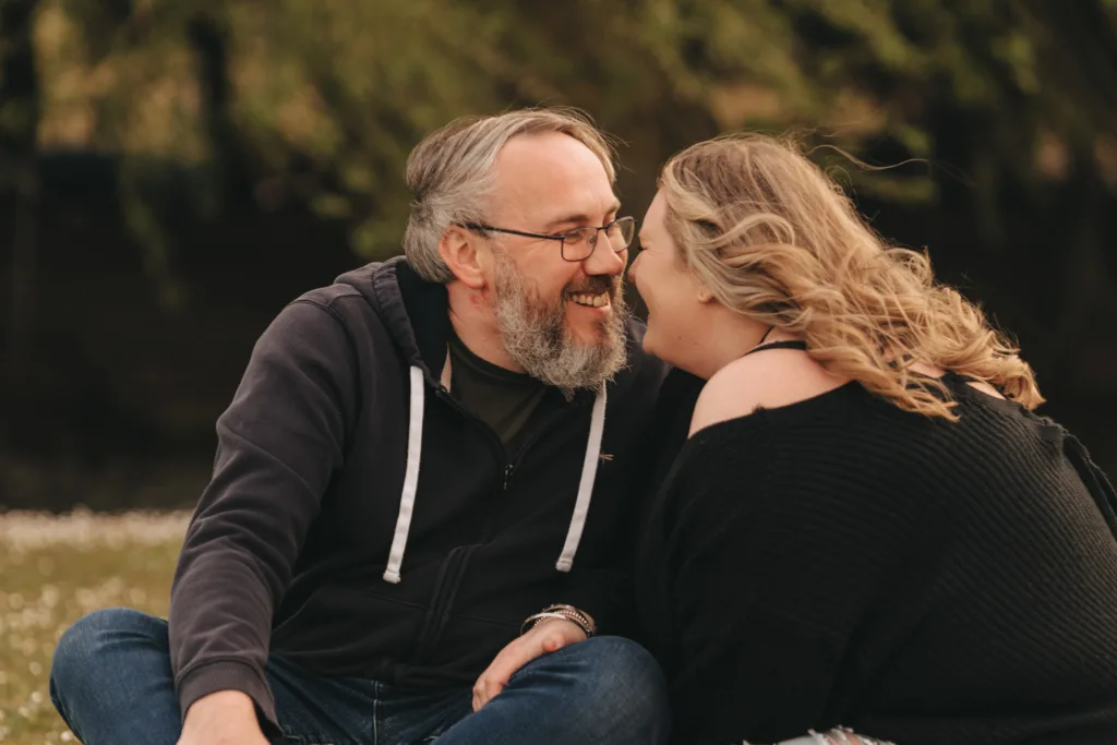 A couple sits closely on the grass by Cleethorpes Boating Lake, smiling at each other. The man, with a gray beard and glasses, wears a black hoodie. The woman, her long wavy blonde hair cascading over one shoulder, sports a black top. Trees in the dark background hint at their engagement moment. © Aimee Lince Photography - Wedding photographer in Lincolnshire, Yorkshire & Nottinghamshire