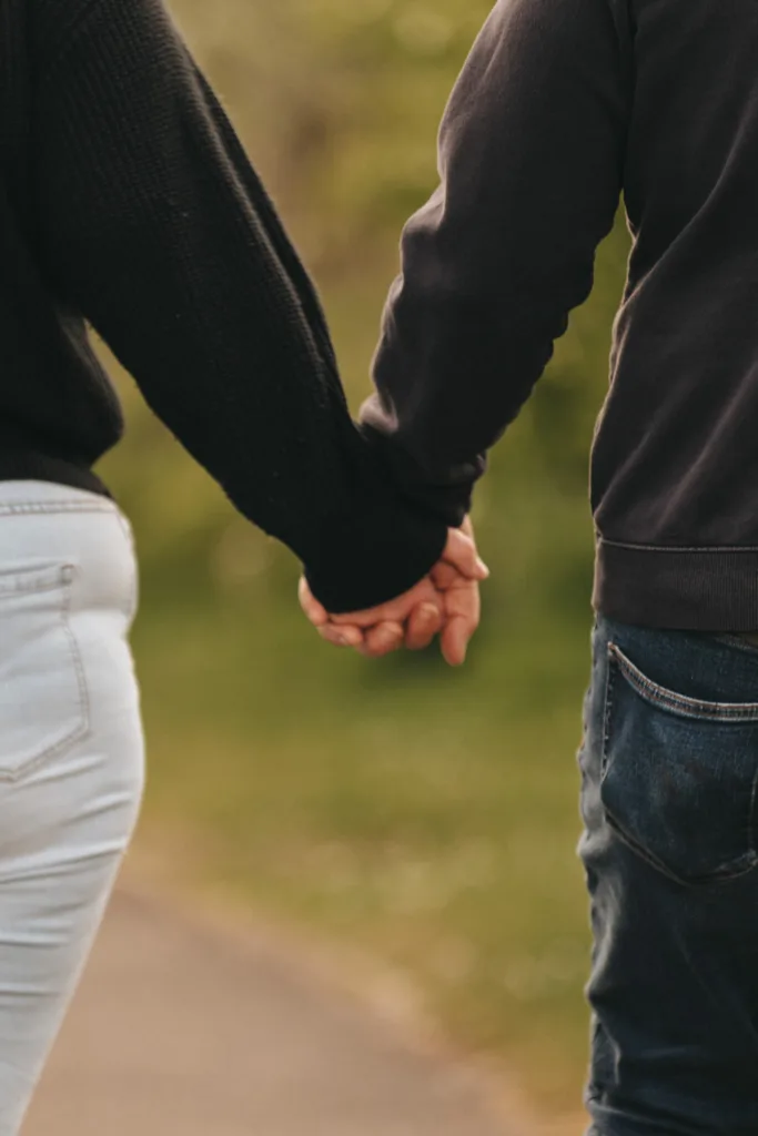 Two people are holding hands, perhaps celebrating their engagement, as they stroll through what seems like a park or garden. The person on the left wears light blue jeans and a dark sweater, while the other dons dark blue jeans and a black sweater. Blurred greenery in the background evokes memories of Cleethorpes Boating Lake. © Aimee Lince Photography - Wedding photographer in Lincolnshire, Yorkshire & Nottinghamshire