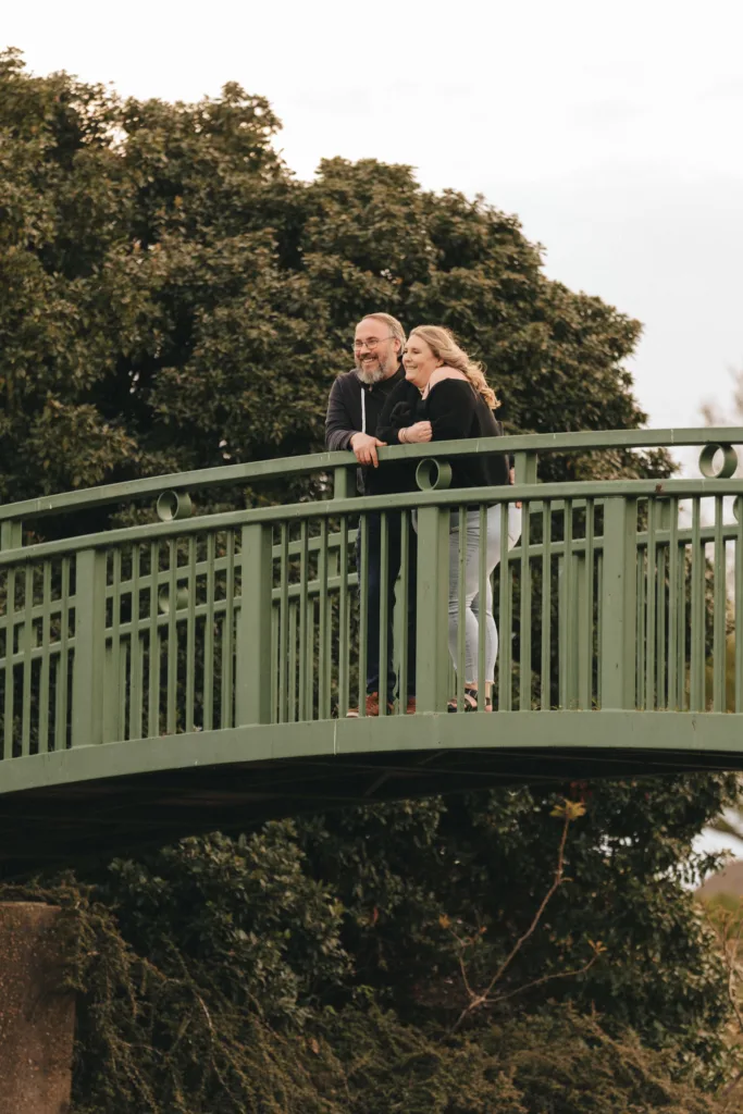 A man and woman stand closely on a green bridge near Cleethorpes Boating Lake, leaning on the railing and smiling. Surrounded by lush greenery, the overcast sky creates a serene atmosphere. The woman wears a black top and jeans, the man in a dark jacket and jeans, announcing their engagement with joy. © Aimee Lince Photography - Wedding photographer in Lincolnshire, Yorkshire & Nottinghamshire
