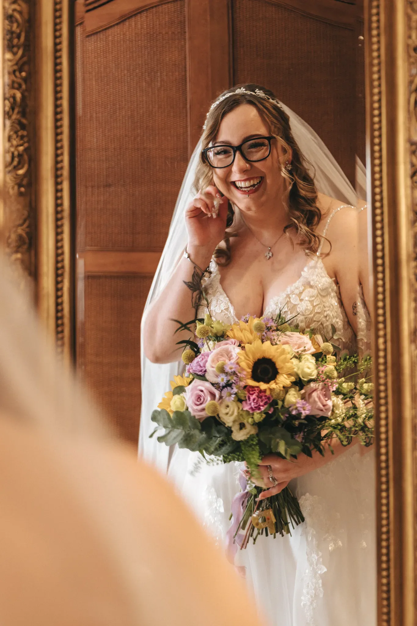 A radiant bride in a lace gown smiles joyfully while adjusting her glasses, clutching a vibrant bouquet of sunflowers and roses, framed by an ornate mirror.