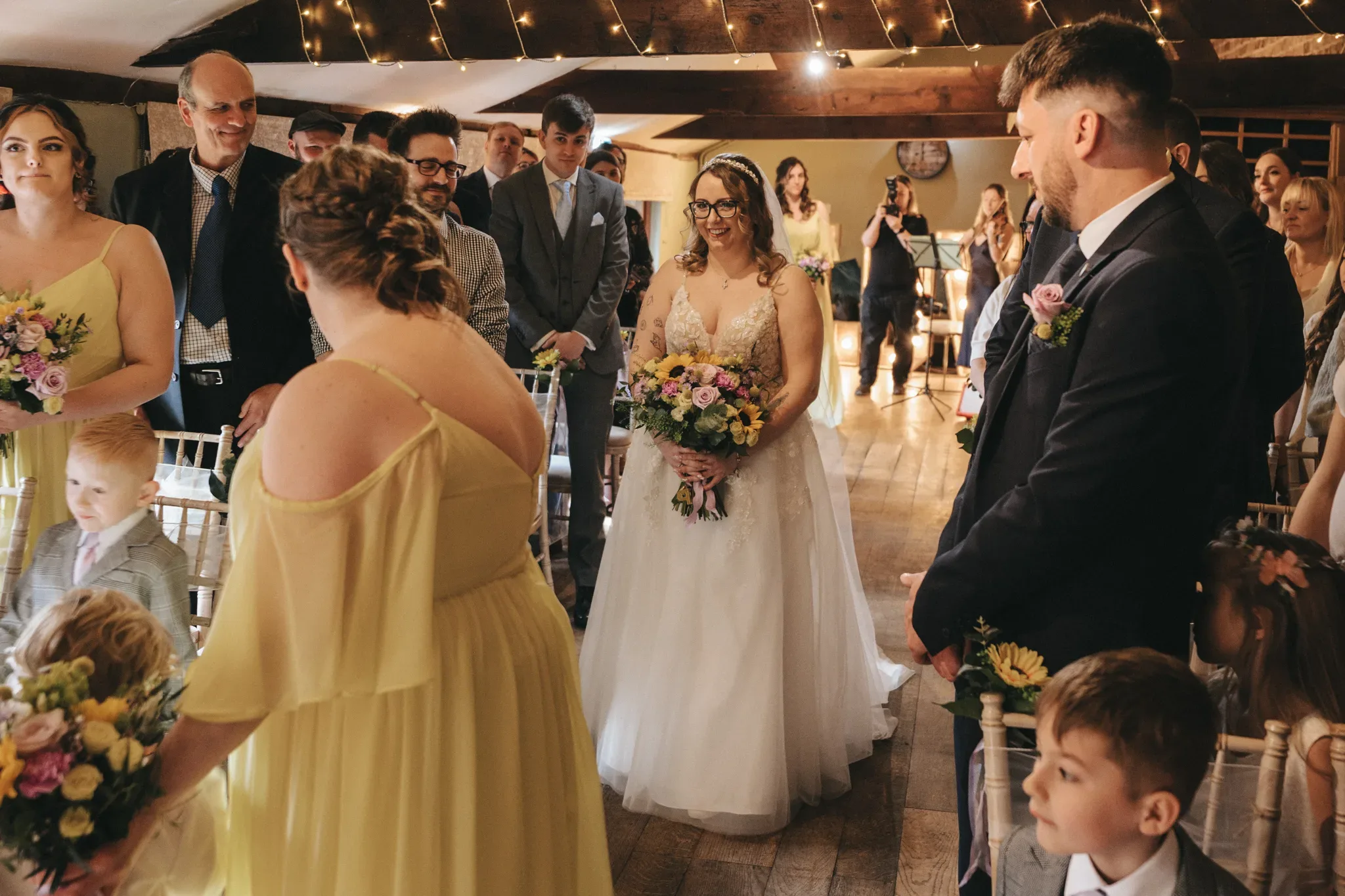 A joyful bride, holding a bouquet, walks down the aisle accompanied by bridesmaids in yellow dresses and a young page boy, as guests and the groom look on fondly in a warmly lit rustic wedding venue.