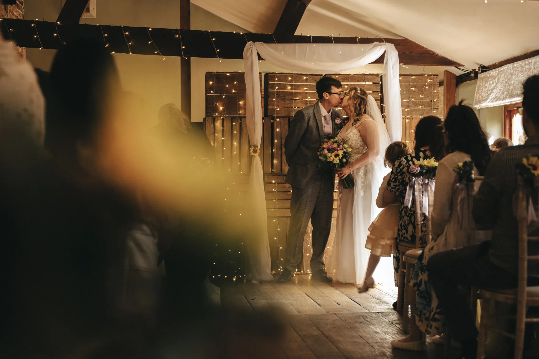 A newlywed couple shares a kiss amidst the warm ambiance of their rustic wedding venue, framed by strings of lights and the affectionate gaze of their guests.