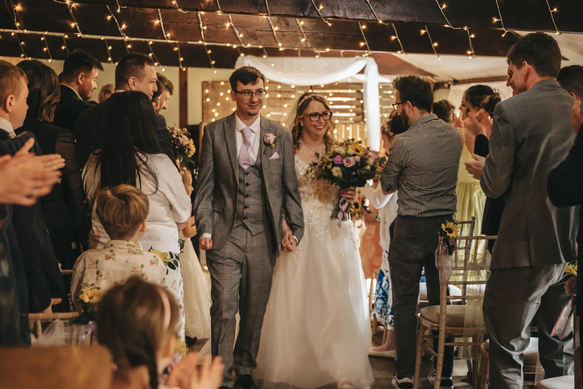 A radiant couple, the bride clutching a lush bouquet, walks joyfully down the aisle among cheering guests in a warmly lit, rustic venue adorned with string lights, marking their first steps as newlyweds.