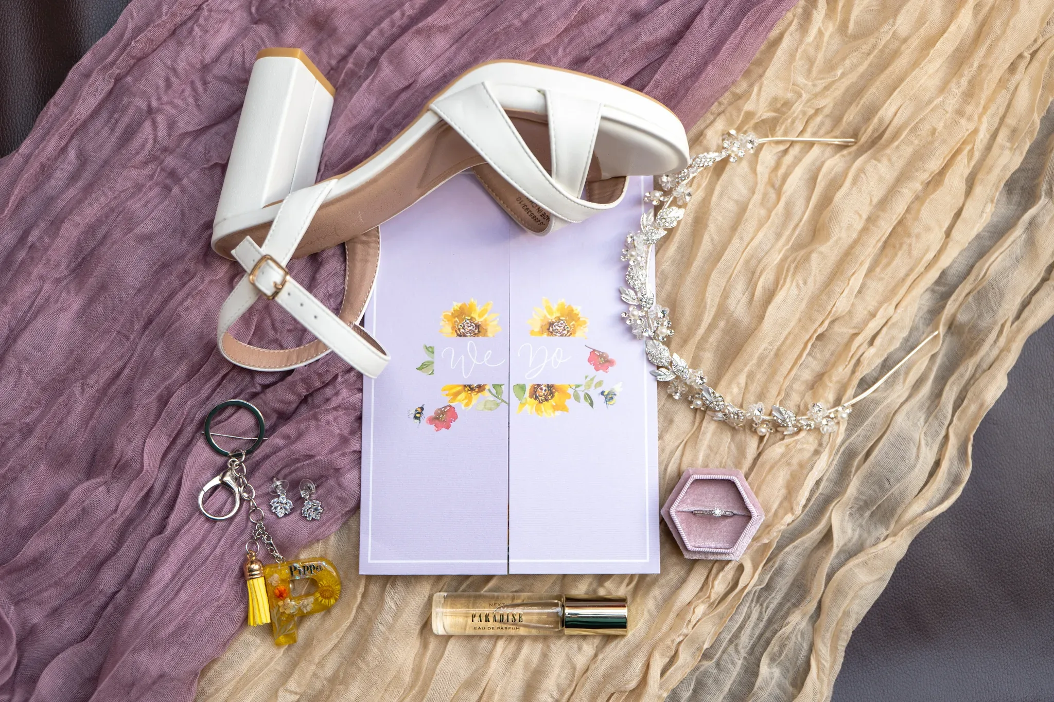 Elegant bridal accessories arranged on a textured surface, featuring white high-heeled shoes, a floral wedding invitation, a crystal necklace, a keychain, and a touch of makeup, all capturing the essence of wedding day preparations.