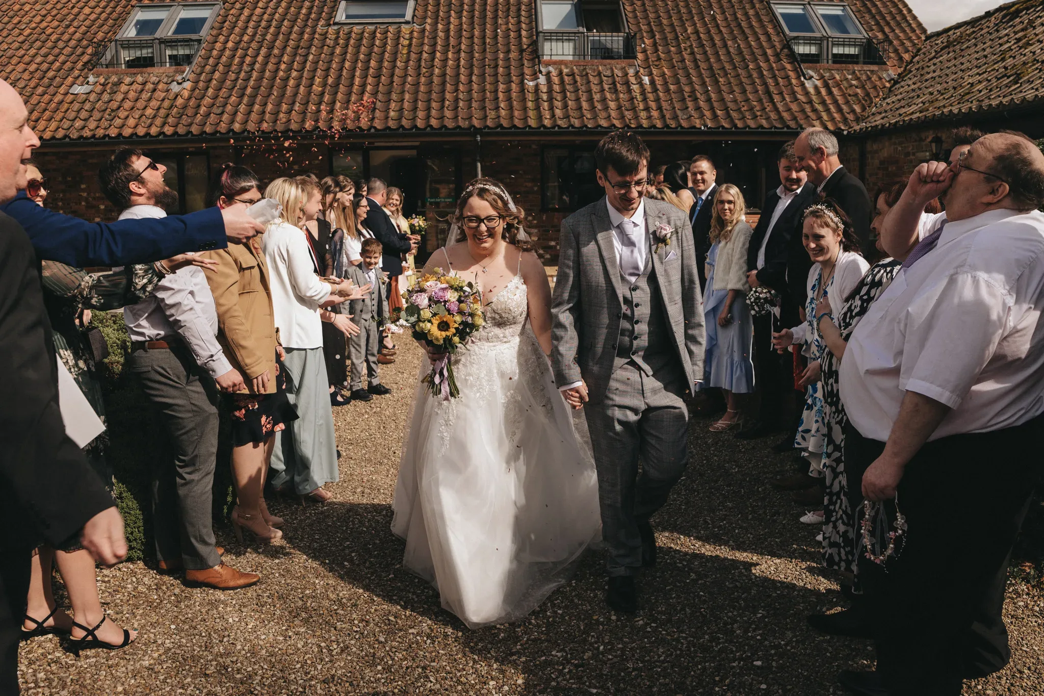 A newlywed couple walks hand-in-hand as guests line up on either side, throwing confetti and cheering to celebrate their marriage, with a rustic brick venue in the background.