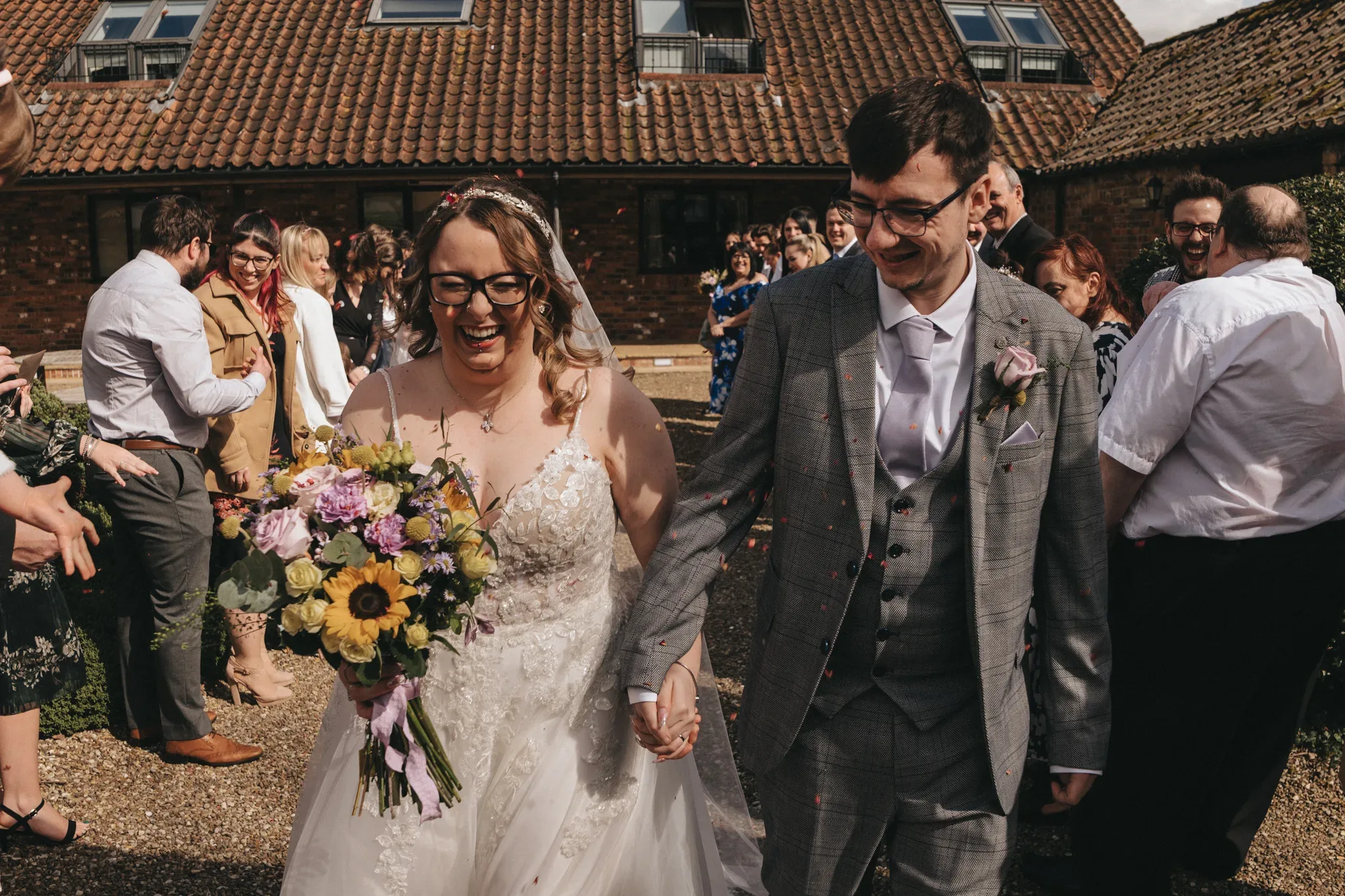 A joyful bride and groom walk hand in hand, smiling widely as they are showered with congratulations from friends and family on a sunny day with a rustic brick building in the background.