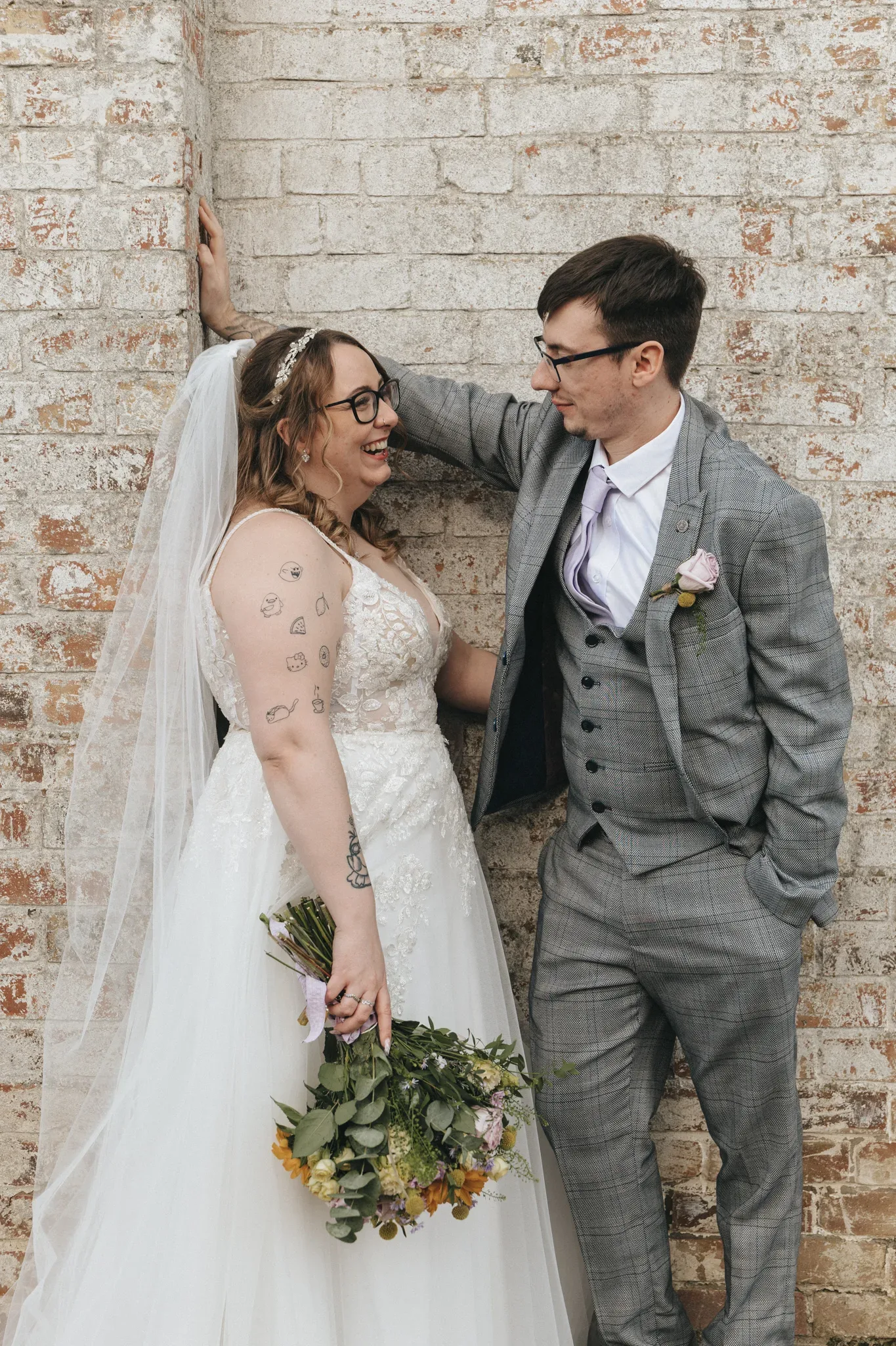 A joyful bride and groom share a candid moment against a brick wall backdrop, the bride holding a bouquet and both dressed in elegant wedding attire.