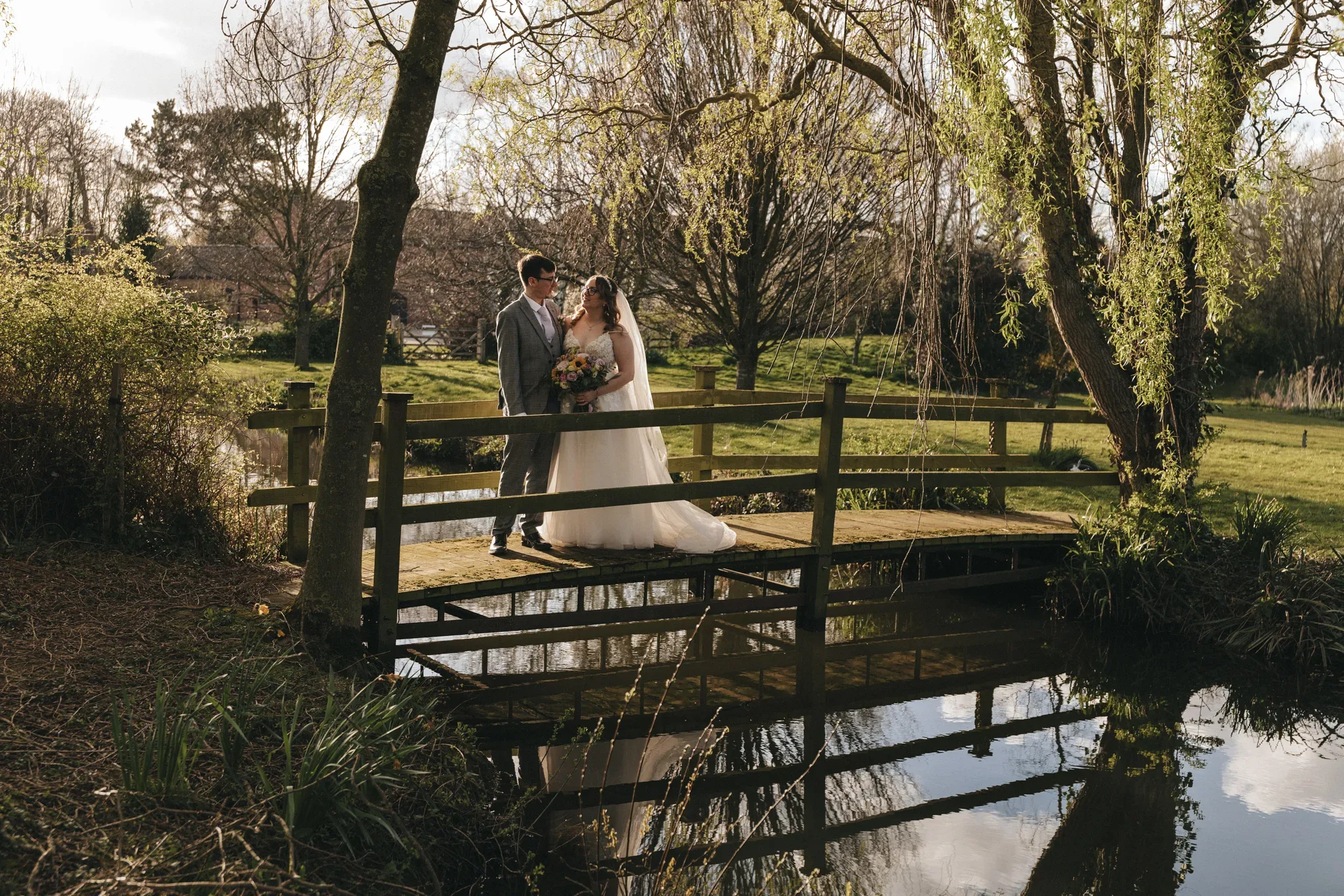 A newlywed couple shares a tender moment on a rustic wooden bridge amidst a serene, sun-dappled landscape, reflecting a picturesque countryside romance.