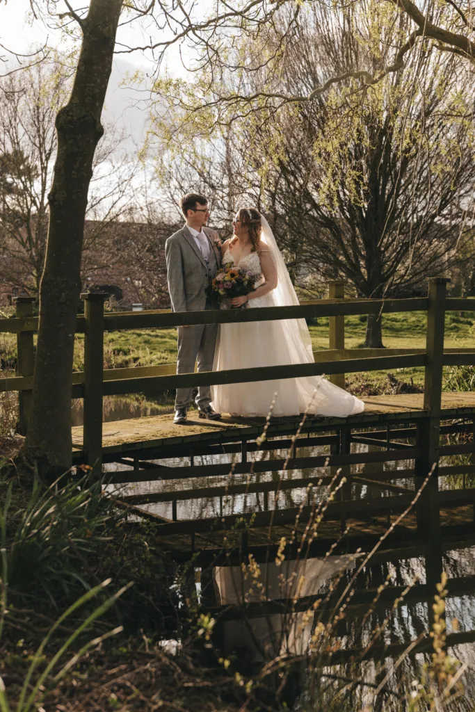 A bride and groom share a tender moment on a wooden bridge amidst a rustic countryside setting, basked in soft sunlight with verdant trees forming a tranquil backdrop.