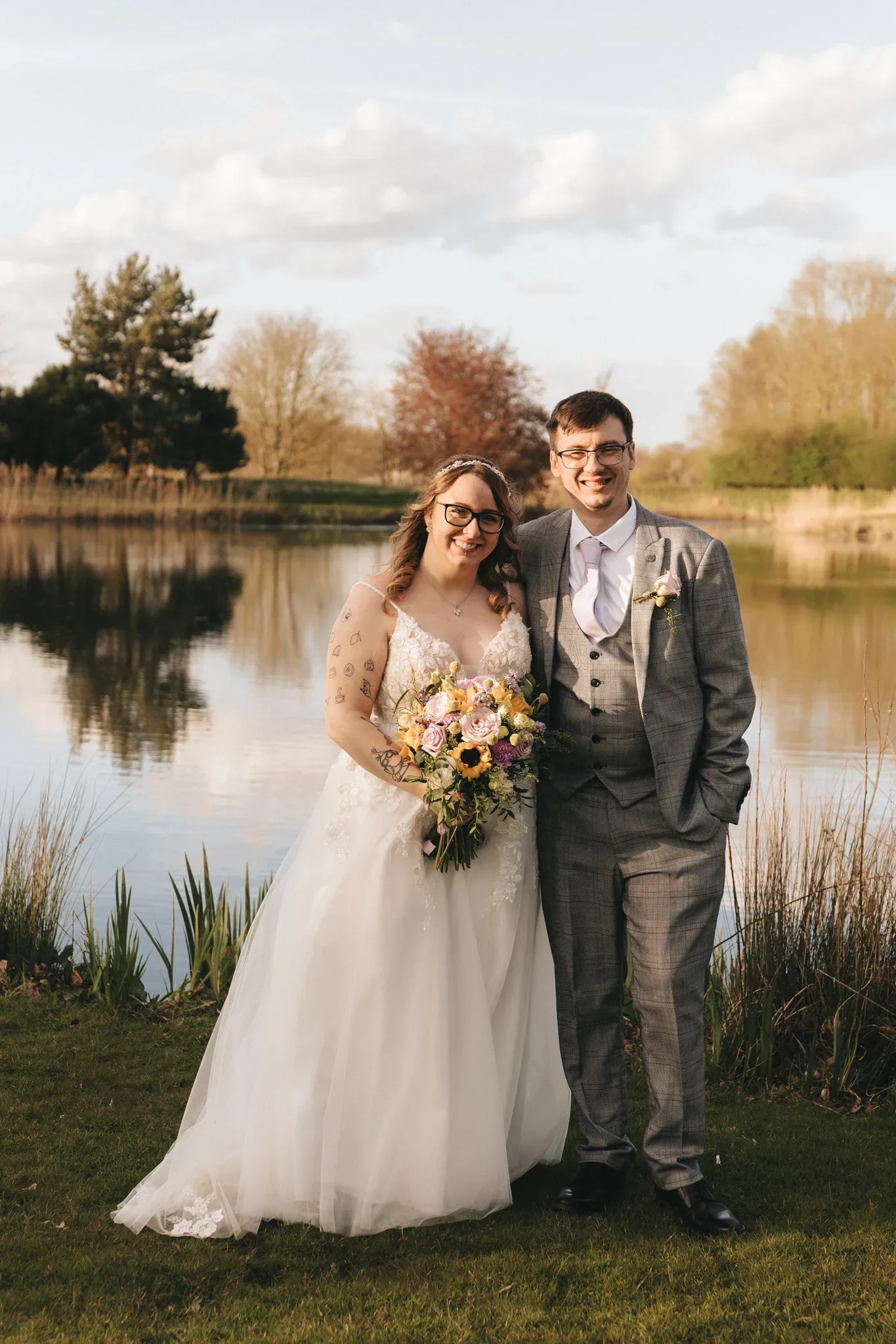 A radiant couple in wedding attire posing with joy by a tranquil lake amidst nature, the bride holding a vibrant bouquet, encapsulating a moment of nuptial bliss.
