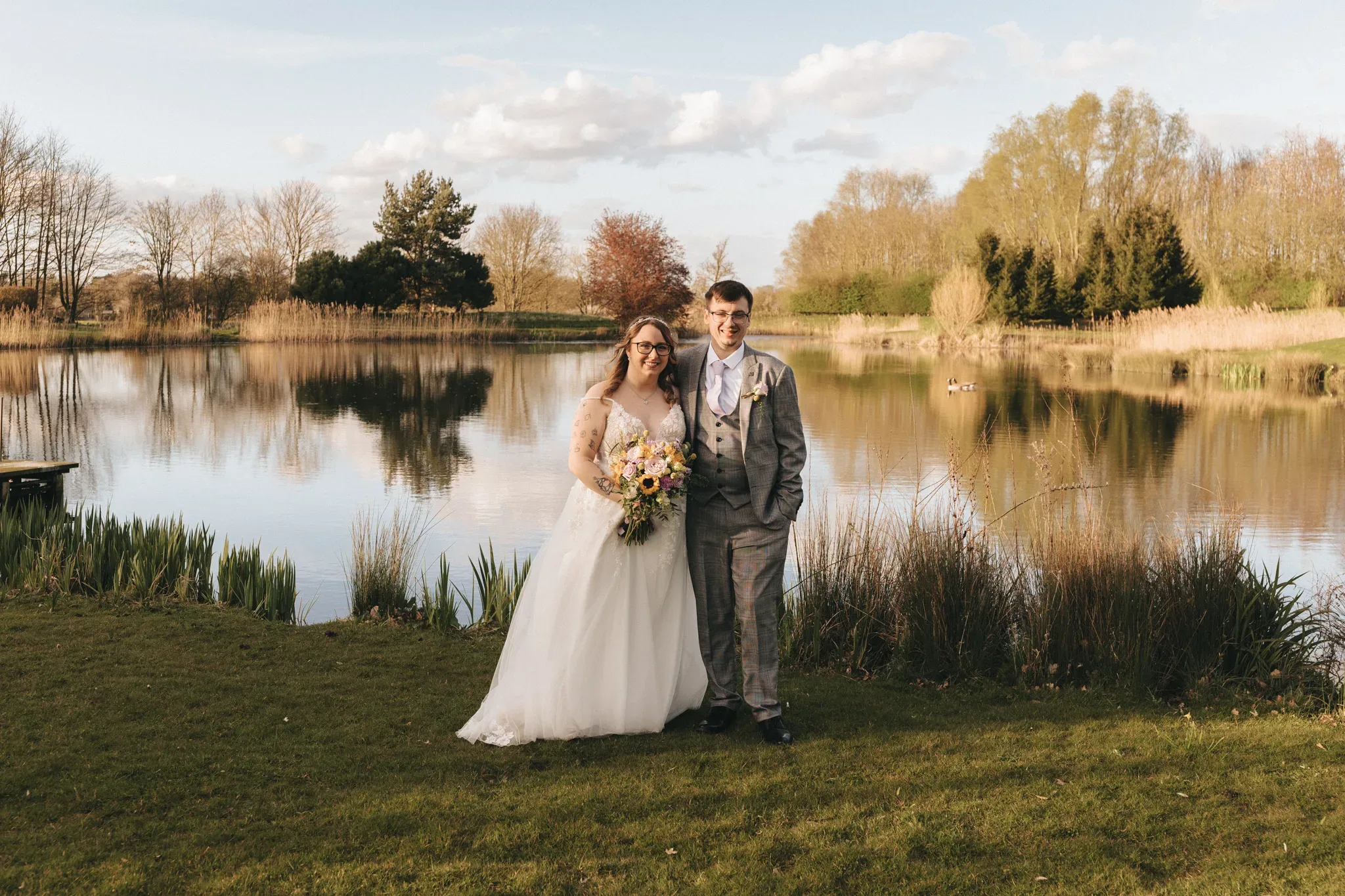A smiling bride and groom stand by a serene pond surrounded by lush greenery under a soft sky, reflecting their joy on their wedding day.