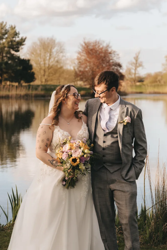 A joyful bride and groom share a tender moment by a tranquil lakeside, bathed in the golden light of a setting sun, with the bride holding a vibrant bouquet of flowers.