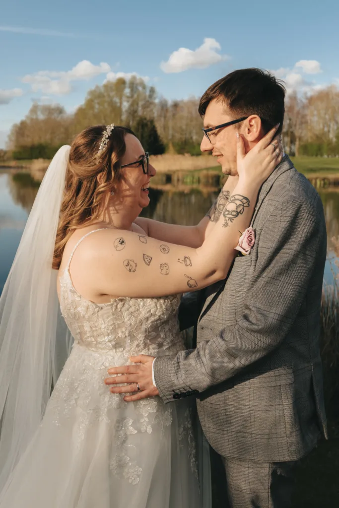 A joyful bride and groom share a tender moment by a tranquil lake, basked in the warm glow of sunlight, their affectionate gaze and smiles speaking volumes of their love.