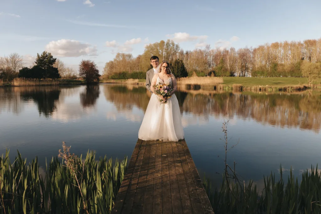 A couple in wedding attire stands on a wooden pier extending into a tranquil lake, surrounded by lush greenery under a soft golden sunset.