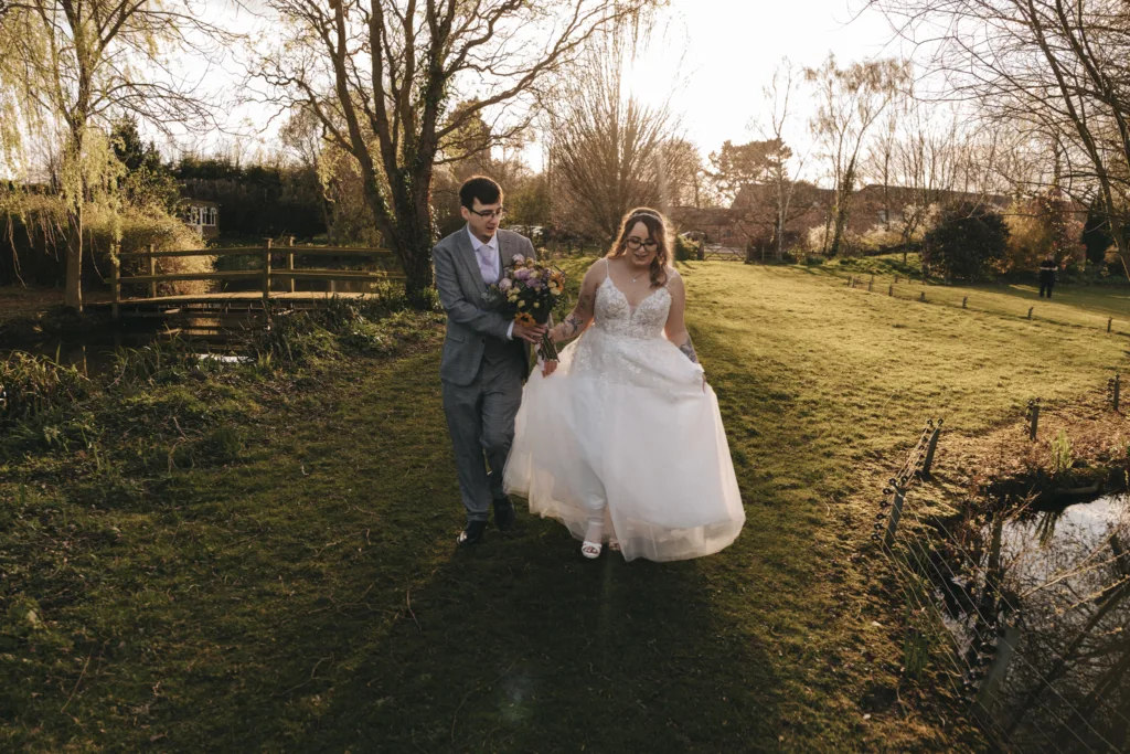 A joyful bride and groom walk hand in hand through a lush garden, with the setting sun casting a warm glow on their wedding day.