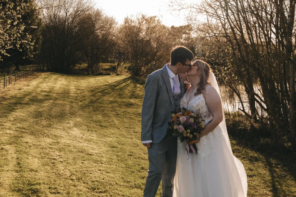 A newlywed couple shares a tender kiss on a sun-drenched meadow, the bride clutching a bouquet of flowers, with serene waters and lush greenery completing the romantic scene.