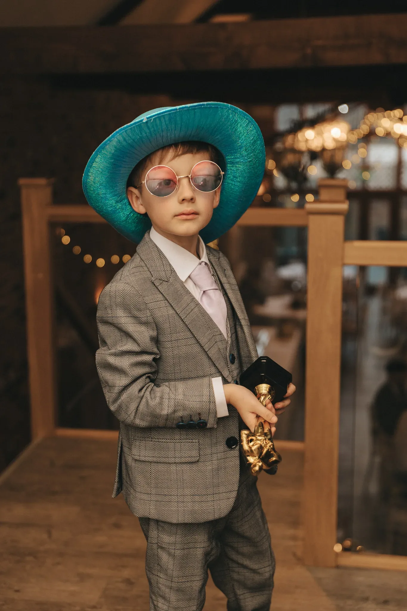 A stylish young boy wearing a gray suit, oversized pink glasses, and a blue hat confidently poses with a golden trophy, exuding a charming and whimsical blend of formality and fun.