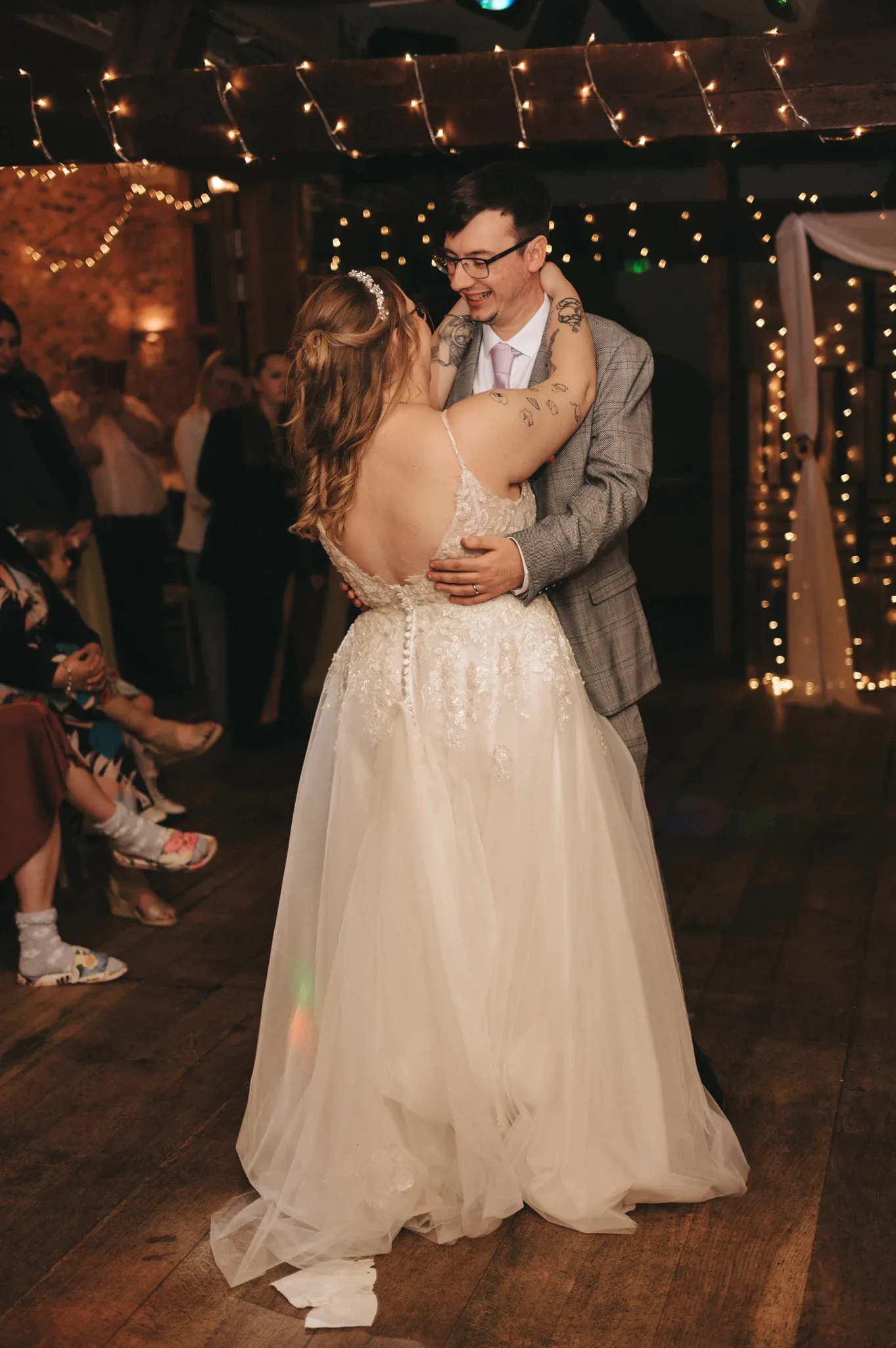 A joyful bride and groom share their first dance under twinkling lights, surrounded by guests, celebrating their wedding in a rustic venue.