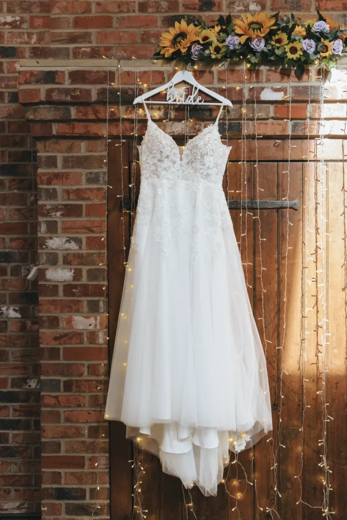 An elegant white bridal gown with lace detailing hangs against a rustic brick wall, adorned with a 'bride' hanger, complemented by twinkling fairy lights and a cheerful garland of sunflowers overhead, capturing the enchanting ambiance of a wedding day.