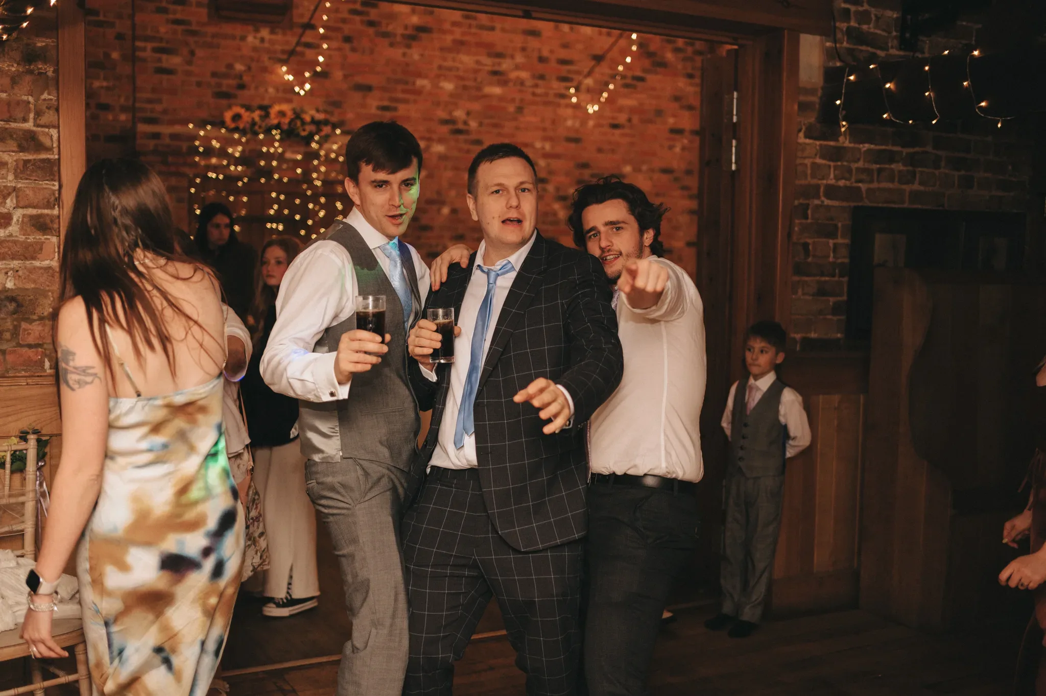 Three men in smart attire enjoy a celebration, sharing a toast with drinks in hand, while lively interactions and a dance floor hint at a wedding or festive party atmosphere.