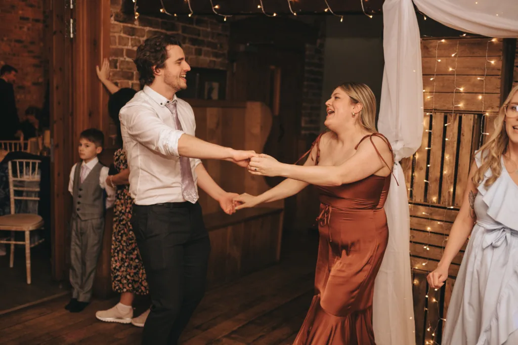 A couple joyfully dances hand-in-hand at an indoor event, likely a wedding, with festoon lighting and onlookers, including a young boy and a woman in the background.