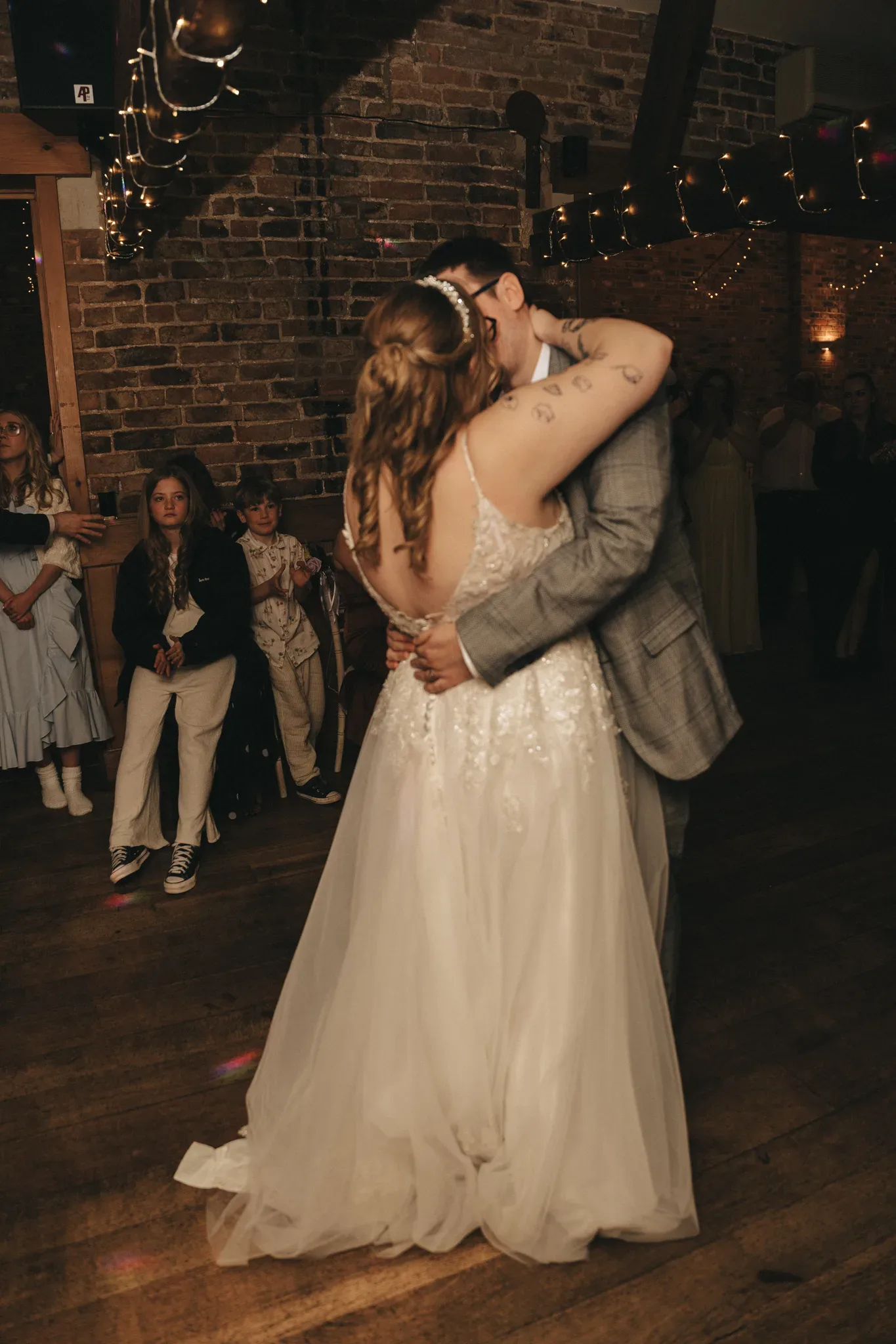 A couple shares their first dance as newlyweds, immersed in a heartfelt moment amidst guests in a warmly-lit rustic venue with exposed brick walls, creating a memory to cherish.