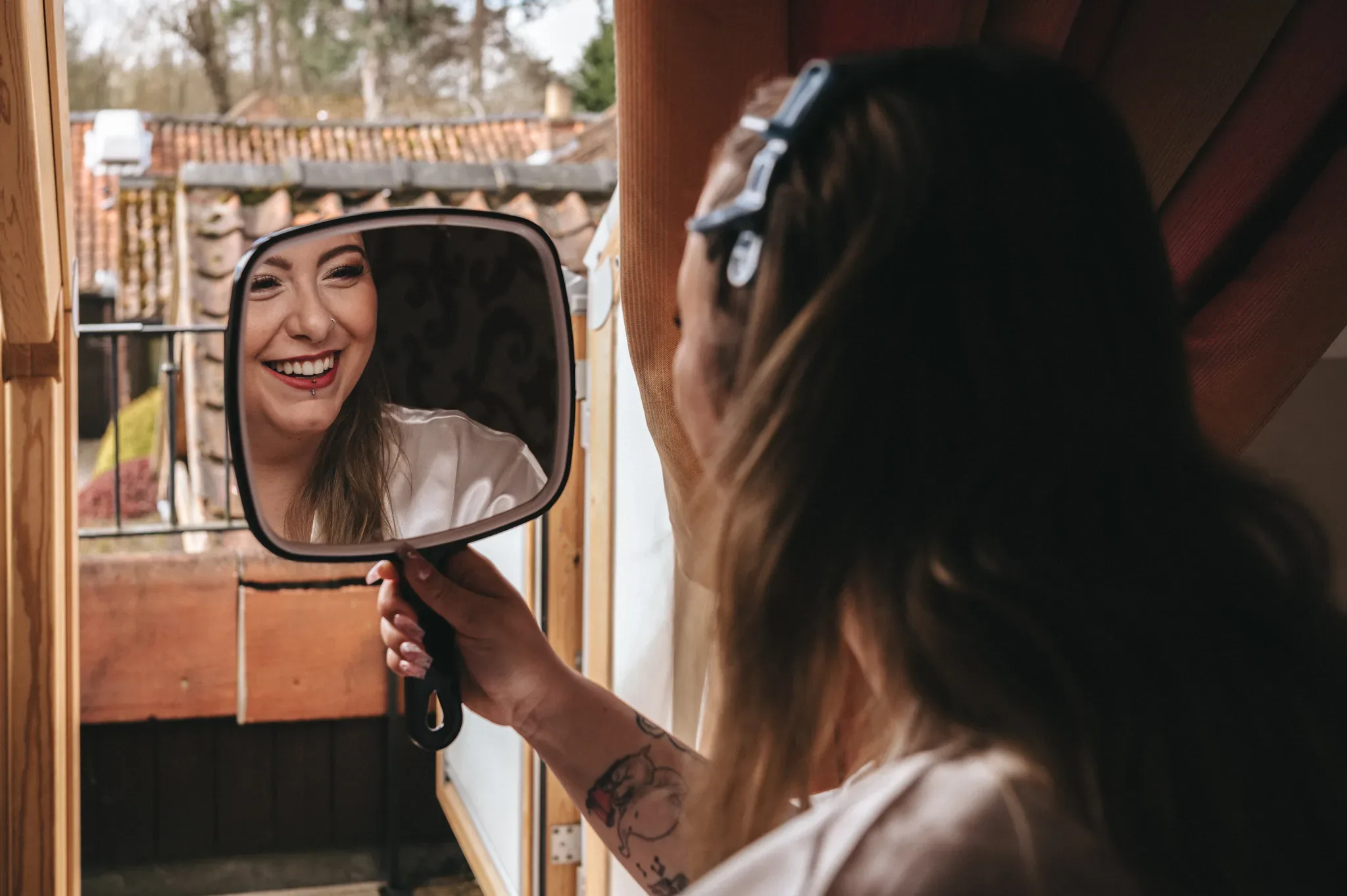 A cheerful woman with sunglasses and a tattoo enjoys her reflection in a hand-held mirror, capturing a moment of happiness and self-appreciation.