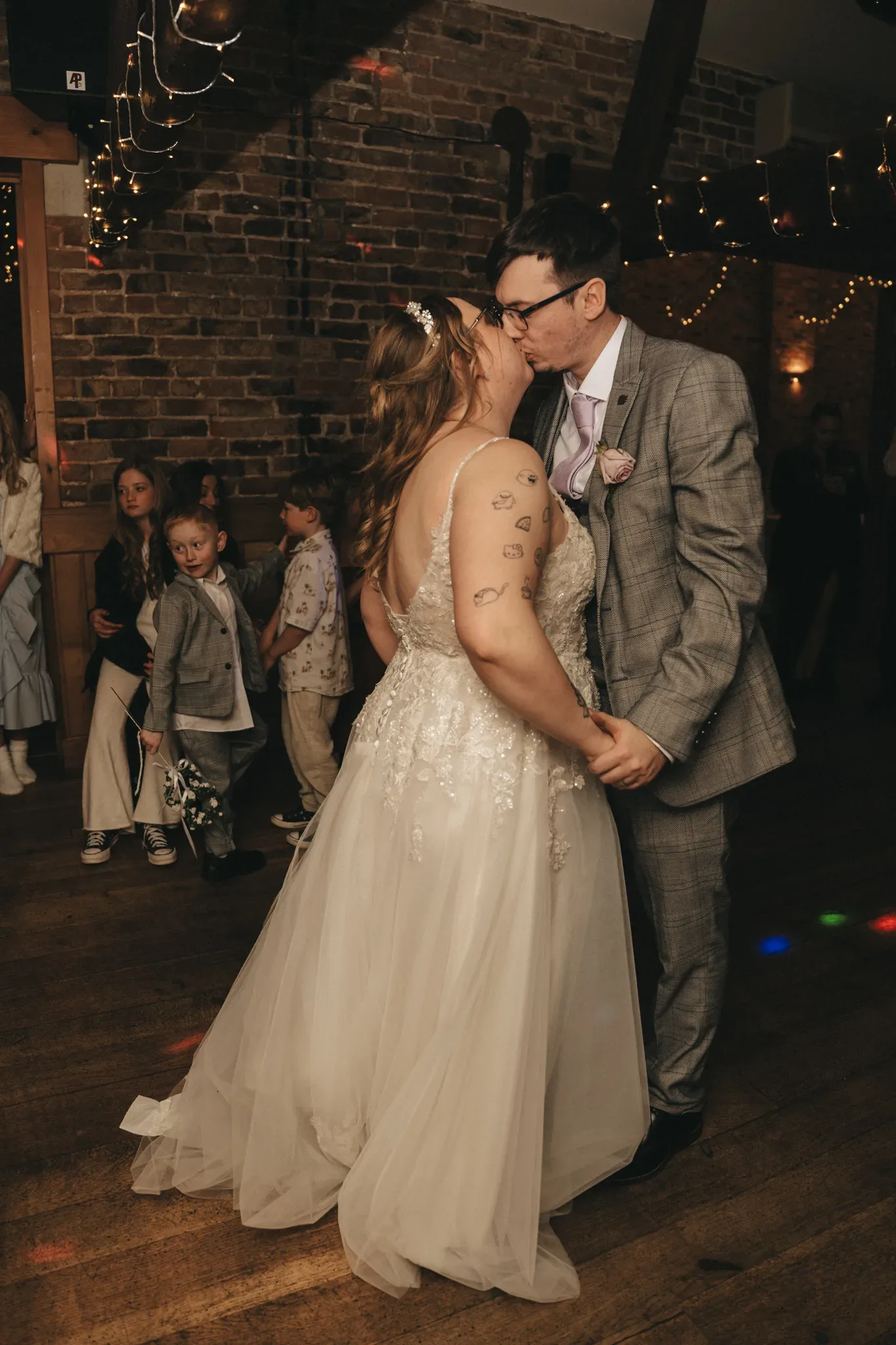 A couple in wedding attire share a romantic dance, their kiss capturing a moment of love, whilst children look on in the warmly lit, rustic reception space.