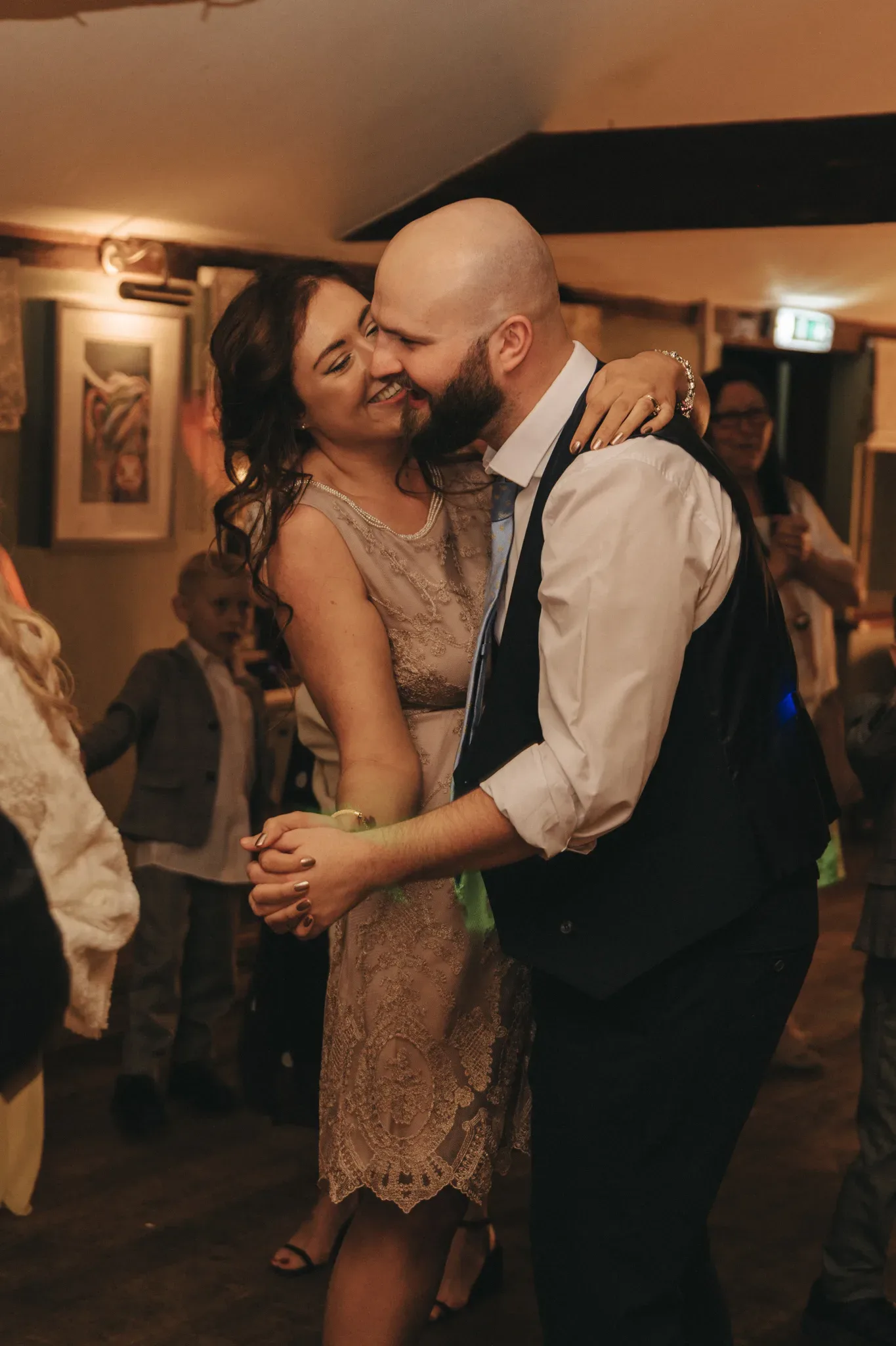 A couple shares a joyous dance, the woman in a lace dress and the man in a vest and tie, both smiling affectionately, surrounded by a warm, celebratory ambiance.