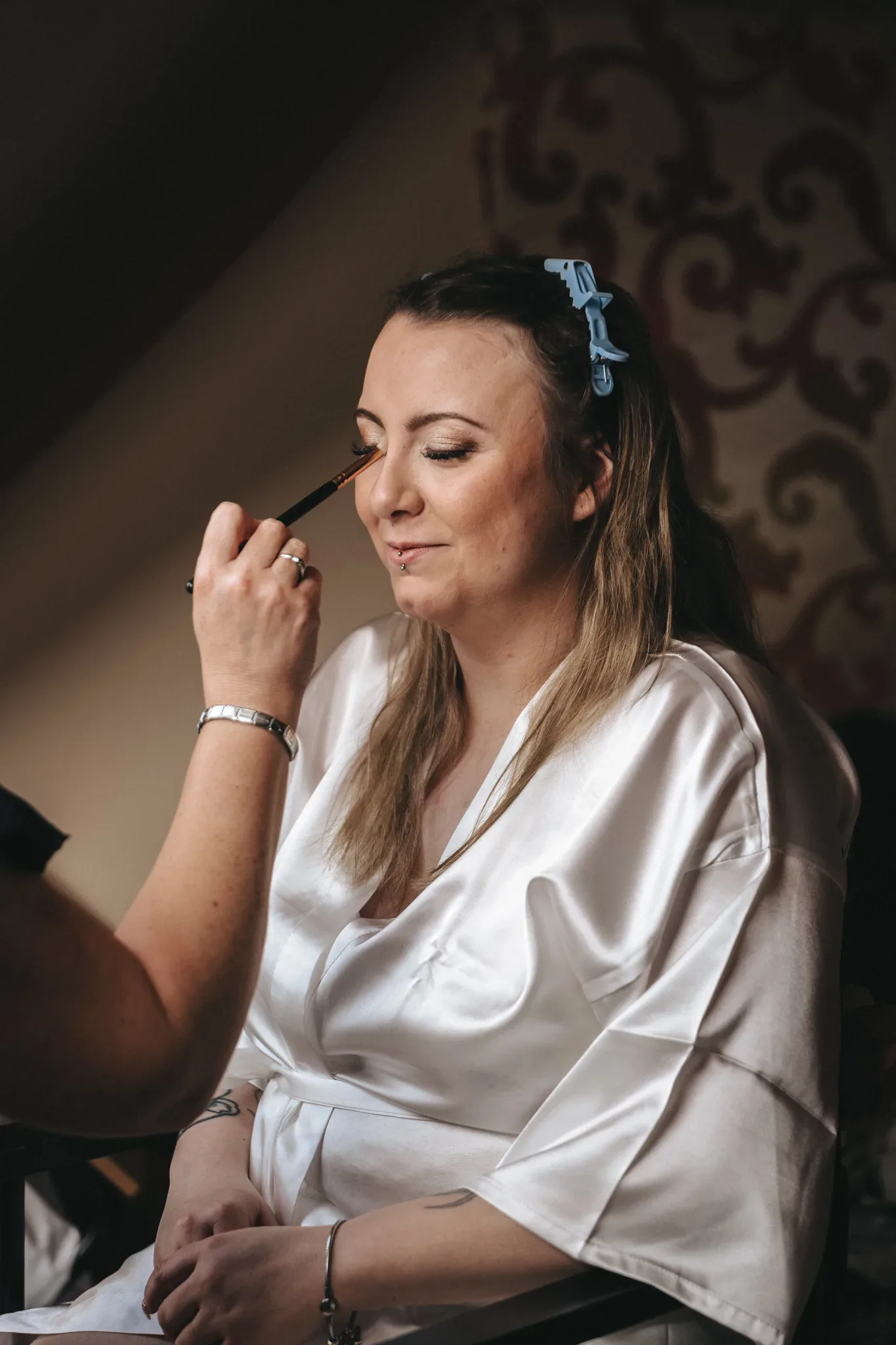 A woman in a white robe sits contentedly as a makeup artist applies eyeshadow, her hair clipped back and a smile hinting on her lips, suggesting a special occasion preparation.