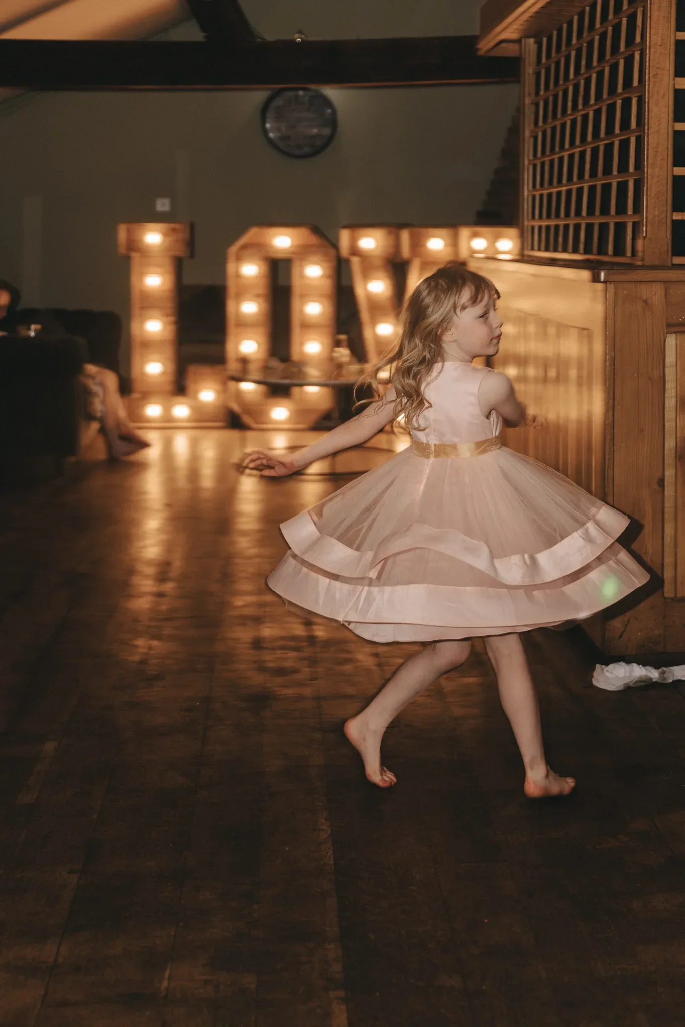 A young girl in a twirling pink dress dances barefoot on a wooden floor, illuminated by the warm glow of large marquee letters in the background.
