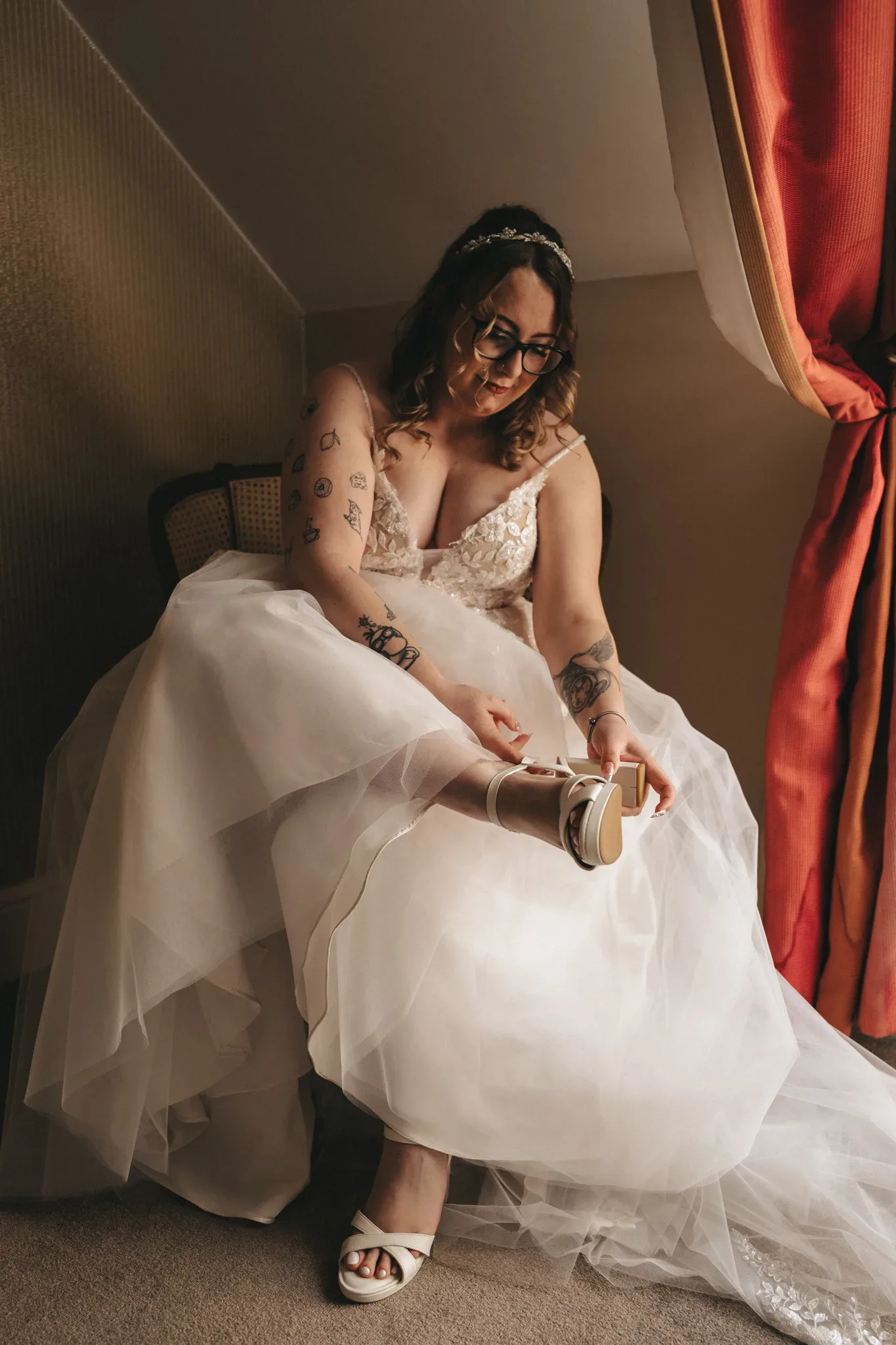 A tattooed bride in a white dress and tiara sits pensively, gently holding her shoe, as soft light filters through a curtain, casting warmth over the scene.