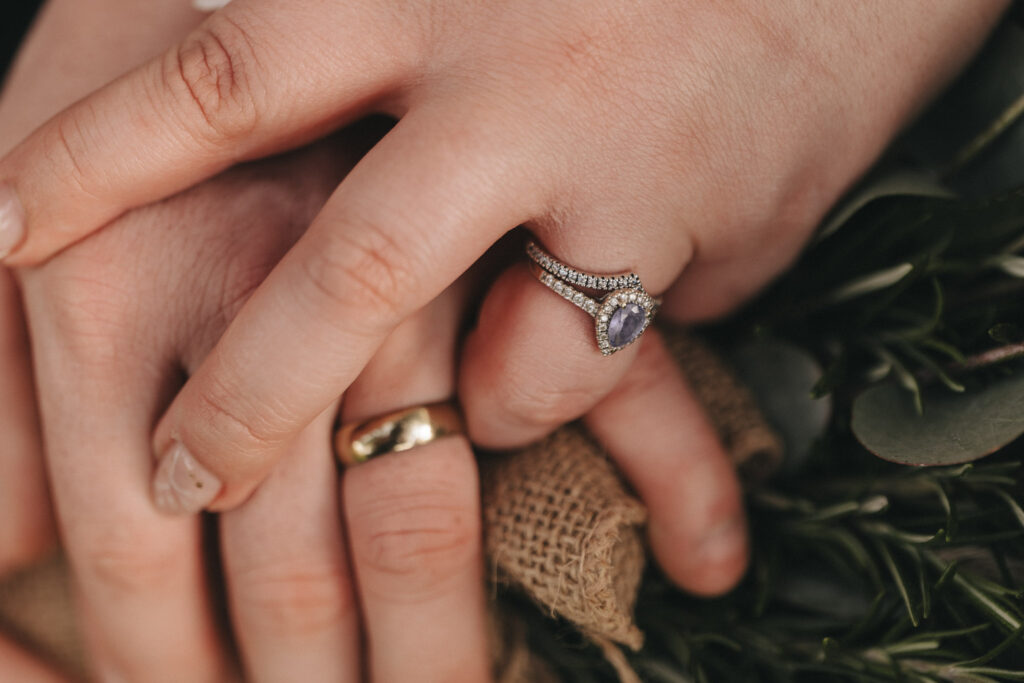 Close-up of two hands clasped together, one wearing a gold band and the other adorned with a sparkling ring featuring a large purple gemstone and smaller surrounding diamonds. The hands, perhaps in the serene setting of Kenwick Park, rest on a burlap-wrapped bouquet with dark green leaves visible in the background. © Aimee Lince Photography