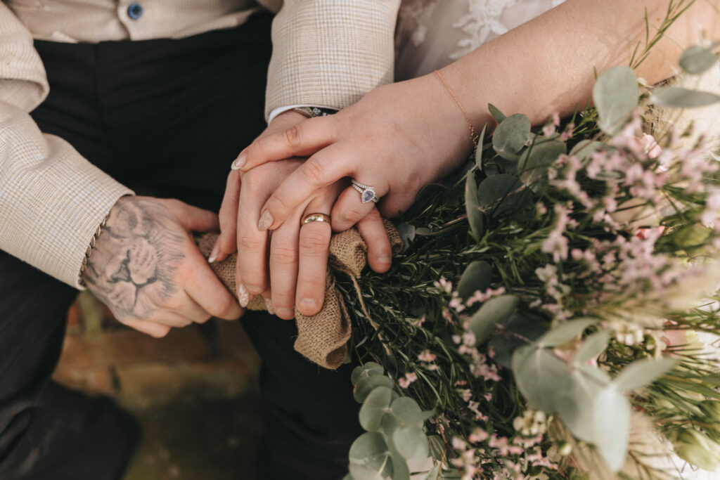 A couple holds hands, showcasing their wedding rings at Kenwick Park. The person on the left, sporting a hand tattoo, wears a light-colored jacket and dark pants. Meanwhile, the person on the right cradles a bouquet of pink and green flowers, wearing a bracelet. Their hands rest gently on a burlap wrap. © Aimee Lince Photography