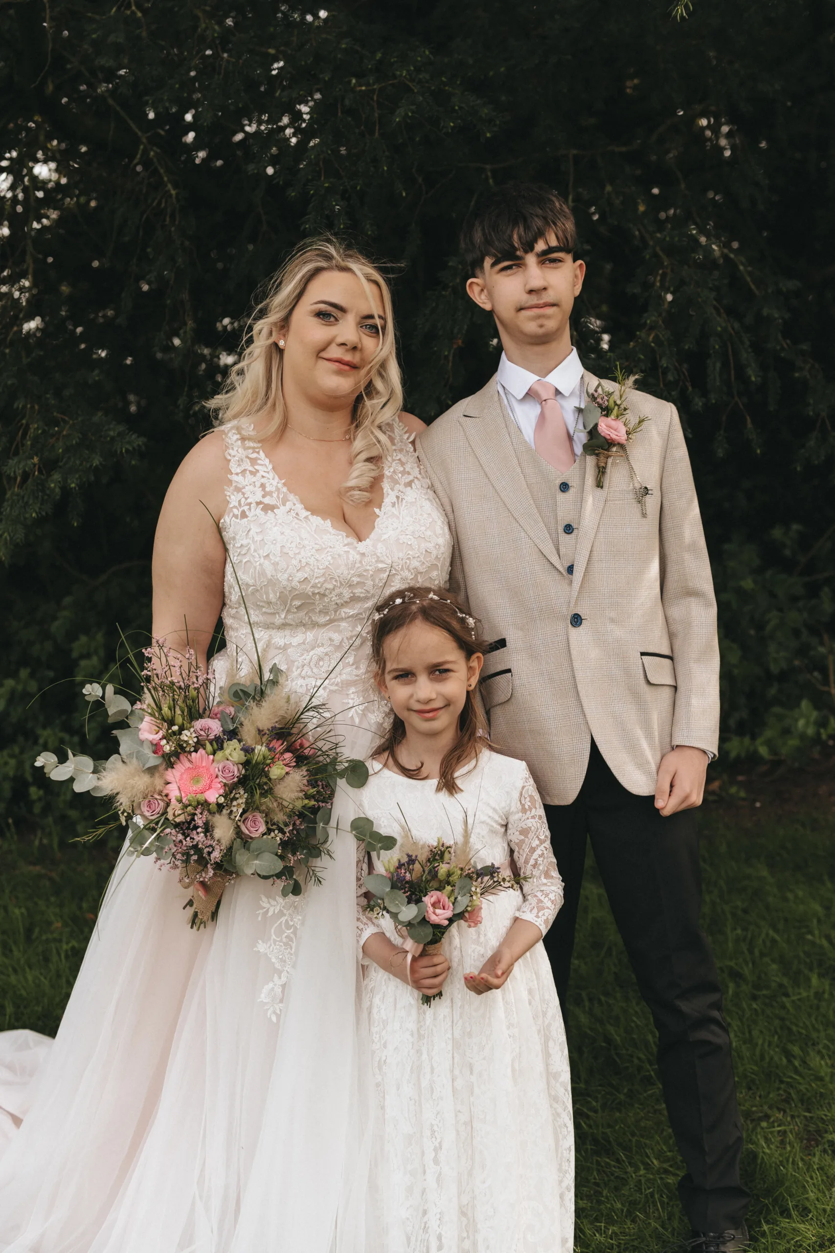A bride named Pippa, a young girl, and teenage boy named Dan pose outdoors. Pippa wears a white lace gown and holds a large bouquet. Dan sports a light gray suit and