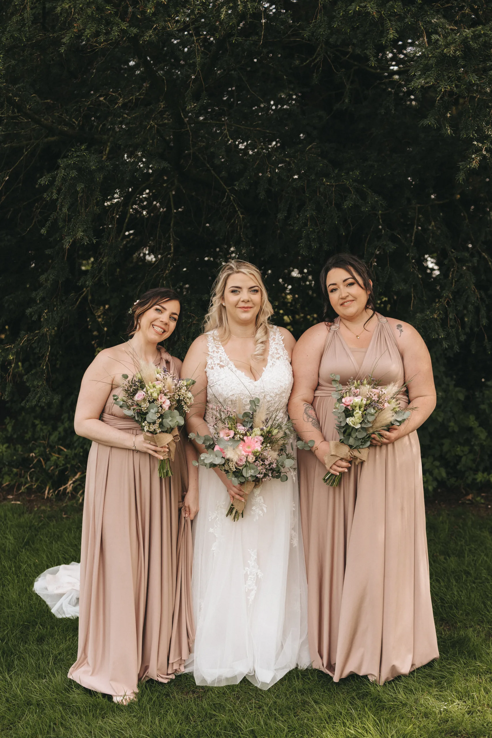 A bride in a white gown stands center among two bridesmaids in blush dresses, all holding bouquets, with a background of lush greenery. The trio smiles joyfully for the camera, capturing the