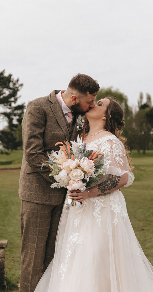 A bride and groom share a kiss outdoors at Grimsby Golf Club. The groom wears a brown suit, and the bride is in a white lace wedding dress, holding a bouquet of flowers. They stand on a grassy field with trees in the background, under a cloudy sky—the perfect scene for stunning photography. © Aimee Lince Photography