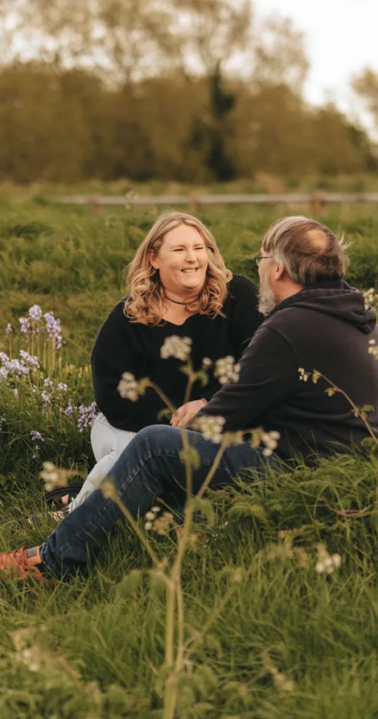 A smiling woman and man sit on the grass in a field near Cleethorpes Boating Lake. The woman wears a black sweater and jeans, while the man sports a dark hoodie and jeans. Blue flowers are nearby, with trees and a rustic fence in the background, creating a warm, engaged atmosphere as they converse. © Aimee Lince Photography - Wedding photographer in Lincolnshire, Yorkshire & Nottinghamshire